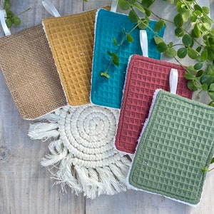 Puede incluir: Un conjunto de esponjas de limpieza coloridas con textura de gofre, cada una con ribete blanco y lazo para colgar. Los colores incluyen marrón, amarillo, turquesa, rojo y verde, dispuestas sobre un posavasos de macramé y una superficie de madera.