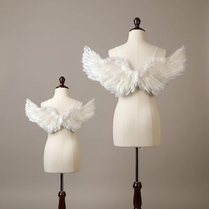 Puede incluir: Dos pares de alas de plumas blancas exhibidas en dos torsos de maniquí de diferentes tamaños. Las alas están construidas con plumas blancas en capas, extendiéndose desde la espalda de cada maniquí, aseguradas con tirantes blancos finos.