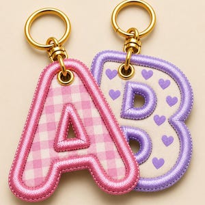 May include: Two embroidered letter keychains with gold-tone metal rings. The letter A features a pink and white gingham pattern with a pink stitched border. The letter B is white with purple hearts and a purple stitched border. Text reads Embroidery Club.