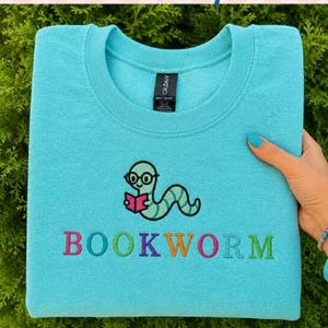 May include: A bright turquoise crewneck sweatshirt featuring an embroidered design of a light green worm wearing glasses and reading a pink book. Below the worm, the word BOOKWORM is embroidered in multicolored letters. The label reads GILDAN.