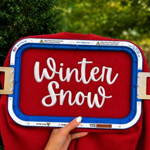 May include: A red sweatshirt secured in a blue rectangular embroidery hoop. The fabric features white embroidered cursive text that reads Winter Snow. The hoop frame includes safety warning labels and text indicating it is a 14 cm Mighty Hoop.