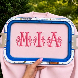 May include: A light pink sweatshirt secured in a blue 14 cm Mighty Hoop embroidery frame. The fabric features a centred, dark pink embroidered monogram of the letters M, I, and A, each flanked by decorative bow accents in matching thread.