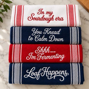 May include: A stack of four rectangular kitchen towels with striped borders. The top white towel reads In my Sourdough era in red. The navy towel reads You Knead to Calm Down, the red towel reads Shhh... I'm Fermenting, and the bottom navy towel reads Loaf Happens.