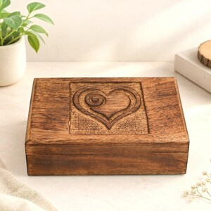 May include: A rectangular wooden box with a dark brown finish and a visible wood grain. The lid features a centered, square-shaped carved panel displaying a stylized heart design with a spiral detail in the center and a textured background.