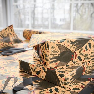 May include: Wrapping paper with a repeating pattern of black bats featuring red eyes, green pine branches, and red berries on a light tan background. The paper is folded over rectangular boxes, with a black ribbon and black scissors nearby.