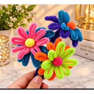 May include: Three handcrafted flowers made from colorful pipe cleaners. One flower has bright pink petals with a yellow center, one is blue with an orange center, and one is lime green and orange with a hot pink center.