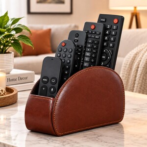 May include: A brown leather remote control holder with a semi-circular side profile and visible stitching. The organizer contains four black remote controls of varying heights, each featuring a numeric keypad, power button, and directional navigation.