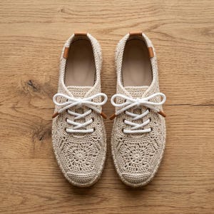 以下が含まれることがあります： ベージュのかぎ針編みスニーカー一足。花をモチーフにした編み込み模様、白い靴紐、かかと部分のタンカラーのレザー調アクセントが特徴。丸みを帯びた爪先と、織り生地を使用したローカットのシルエットが施されています。