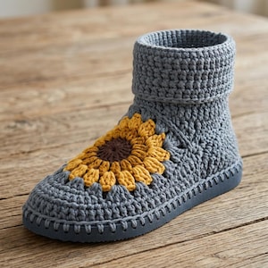 Könnte beinhalten: Ein grauer Häkel-Stiefel mit einem Sonnenblumenmotiv auf der Oberseite. Die Sonnenblume hat eine dunkelbraune Mitte, die von leuchtend gelben Blütenblättern umgeben ist. Der Stiefel hat einen dicken Umschlag und eine graue Sohle.