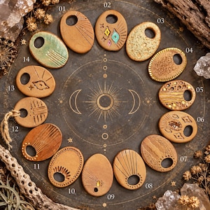 May include: Fourteen oval-shaped ceramic pendants arranged in a circle on a dark surface. Each pendant features unique etched or painted designs, including sunbursts, geometric shapes, and textures, numbered 01 through 14.