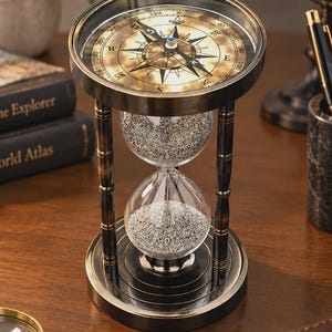 Puede incluir: Un reloj de arena decorativo con marco de metal color latón y una brújula integrada en la parte superior. Los bulbos de vidrio transparente contienen arena fina de color plateado. La esfera muestra los puntos cardinales.