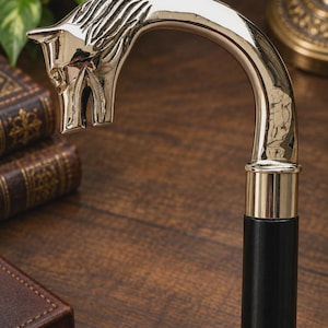 May include: A walking stick featuring a polished gold-tone metal handle shaped like a horse head with a curved neck. The handle connects to a smooth, cylindrical black wooden shaft via a simple gold-tone metal collar, creating a sturdy design.