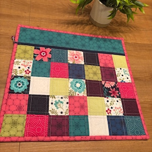 May include: A square quilted fabric pouch featuring a patchwork design of various squares in shades of teal, lime green, dark purple, and hot pink. The pouch includes a dark blue zipper closure along the top edge and a bright pink fabric border.