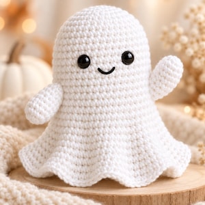 May include: A white crocheted ghost plush toy featuring a rounded head, two small black plastic safety eyes, and a stitched black curved mouth. The ghost has two small arms, one of which is raised in a waving gesture, and a flared base.