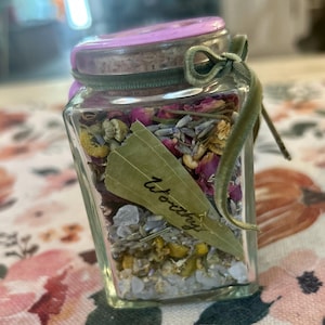 May include: A square glass jar with a pink lid and a green velvet ribbon tied around the neck. The jar is filled with dried flowers, herbs, and small clear crystals. A single dried bay leaf with the word Worthy written on it is inside.