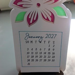 May include: A layered paper desk calendar for January 2027 featuring a white, pink, and green floral cutout design at the top. The calendar is held upright by two small brown wooden stands and displays the month, year, and days of the month in black.