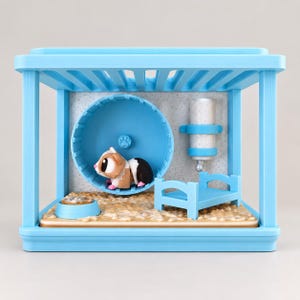 Peut inclure: Un ensemble de jeu pour hamster en plastique bleu clair comprenant une roue d'exercice circulaire, un petit biberon, un lit miniature et une gamelle. Une figurine de hamster marron, blanc et noir est placée dans la roue d'exercice bleue.
