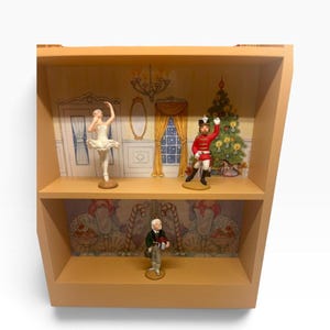 May include: A two-tiered wooden display shelf featuring a light wood finish. The top shelf displays a white ballerina figurine and a red-uniformed soldier figurine against a holiday-themed backdrop. The bottom shelf holds a single male figurine.