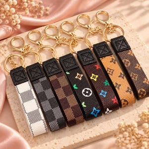 May include: A collection of seven rectangular keychains with gold-toned metal lobster clasps and rings. The straps feature various patterns including white, black, and brown checkerboard designs, as well as black straps with colorful floral motifs.