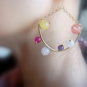 May include: Gold-toned hoop earrings with a chain and colorful gemstone accents. The earrings feature a variety of round gemstones in shades of pink, yellow, purple, and white. The earrings are designed with a unique, curved shape.