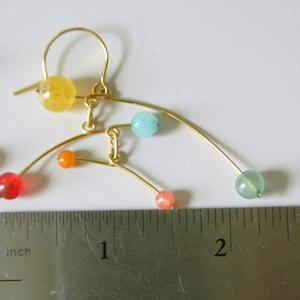 Peut inclure: Boucles d'oreilles en fil dor&eacute; avec des perles de pierres pr&eacute;cieuses color&eacute;es. Les boucles d'oreilles pr&eacute;sentent un design asym&eacute;trique unique avec des perles rouges, oranges, jaunes, bleues et vertes. Les boucles d'oreilles mesurent environ 5 cm de long.
