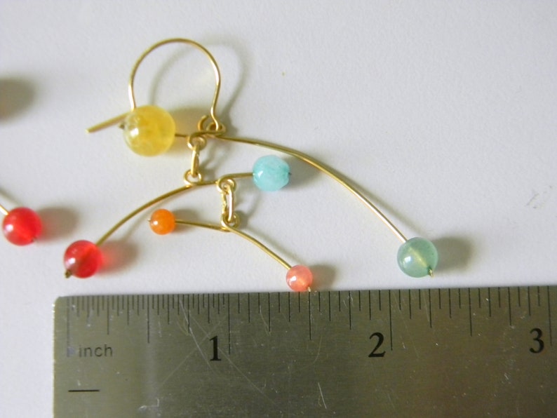 Peut inclure: Boucles d'oreilles en fil dor&eacute; avec des perles de pierres pr&eacute;cieuses color&eacute;es. Les boucles d'oreilles pr&eacute;sentent un design asym&eacute;trique unique avec des perles rouges, oranges, jaunes, bleues et vertes. Les boucles d'oreilles mesurent environ 5 cm de long.