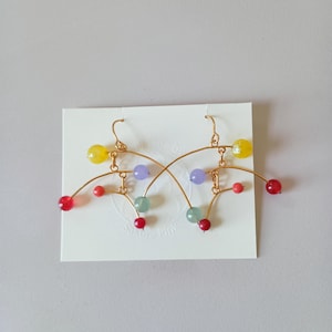 Peut inclure: Une paire de boucles d'oreilles en fil d'or avec des perles color&eacute;es. Les boucles d'oreilles pr&eacute;sentent des perles jaunes, rouges, violettes et vertes. Le design est abstrait et moderne, avec des lignes courbes et des perles pendantes. Les boucles d'oreilles sont pr&eacute;sent&eacute;es sur une carte blanche.
