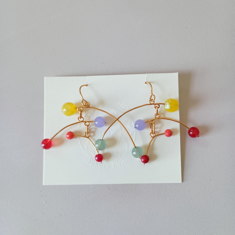Peut inclure: Une paire de boucles d'oreilles en fil d'or avec des perles color&eacute;es. Les boucles d'oreilles pr&eacute;sentent des perles jaunes, rouges, violettes et vertes. Le design est abstrait et moderne, avec des lignes courbes et des perles pendantes. Les boucles d'oreilles sont pr&eacute;sent&eacute;es sur une carte blanche.