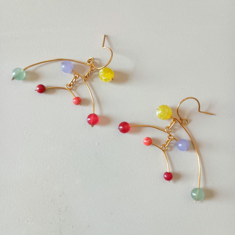 Peut inclure: Une paire de boucles d'oreilles dor&eacute;es au design abstrait unique. Chaque boucle d'oreille pr&eacute;sente un fil incurv&eacute; orn&eacute; de perles color&eacute;es rouges, jaunes, vertes et violettes. Les perles sont de tailles vari&eacute;es, ajoutant de l'int&eacute;r&ecirc;t visuel.