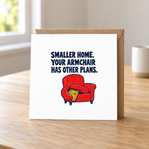 May include: A square greeting card with a white background featuring a red armchair illustration holding a small yellow door. The text reads: SMALLER HOME. YOUR ARMCHAIR HAS OTHER PLANS. The card is placed against a brown envelope on a wooden surface.