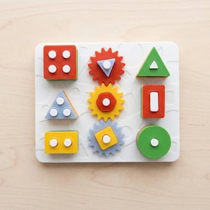 Op de afbeelding: Een wit rechthoekig houten puzzelbord met negen kleurrijke geometrische vormen. De vormen omvatten vierkanten, driehoeken, cirkels en tandwielachtige figuren in rood, geel, groen en blauw, elk met kleine witte pinnetjes om te sorteren.