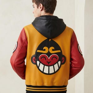 Peut inclure: Une veste style universitaire avec un corps jaune moutarde, des manches rouges et une capuche en cuir noir. Le dos présente un grand motif circulaire représentant un visage de singe souriant en noir, rouge et blanc. Bords-côtes noirs rayés.