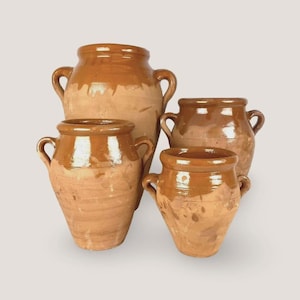 Può includere: Un set di tre vasi in terracotta, ciascuno dotato di due manici. I vasi presentano una base in argilla opaca naturale con una smaltatura lucida color miele che copre la parte superiore e il bordo, creando un distinto aspetto bicolore su ogni vaso.