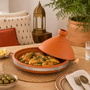 Può includere: Un tajine in terracotta con coperchio conico, caratterizzato da una base larga con bordo in metallo e manici in legno. Il tajine contiene un piatto a base di pezzi di pollo e olive verdi. L'esterno del recipiente in argilla è color arancio.