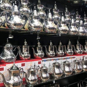 Puede incluir: Una colección de teteras de metal color plata pulida dispuestas en varios estantes. Las teteras cuentan con picos curvos, tapas abovedadas con remates y asas resistentes. Algunas teteras se exhiben sobre sus cajas de producto.