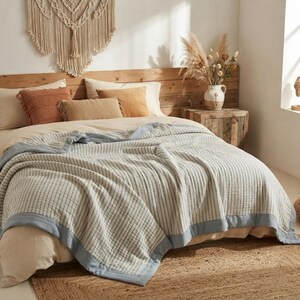 May include: A light blue and white striped cotton blanket with a solid light blue border, draped over a bed with beige and terracotta pillows, a wooden headboard, and a ceramic vase filled with dried pampas grass and flowers.