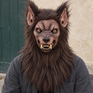 Peut inclure: Un masque de loup-garou couvrant toute la tête, doté d'une fausse fourrure brun foncé, d'oreilles pointues et d'un museau texturé avec des dents acérées visibles. Le masque présente des yeux jaunes perçants et une expression menaçante.