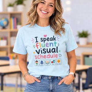 May include: A light blue t-shirt featuring the text "I speak fluent visual schedule" in colorful lettering. Below the text are five small square icons representing "hello," "school," "work," "snack," and "all done" with corresponding illustrations.
