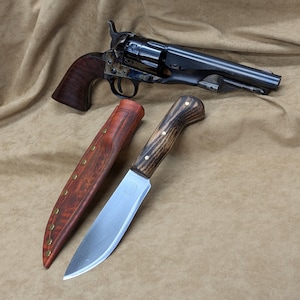 May include: A black revolver with a wooden grip and a stainless steel knife with a wooden handle and a leather sheath. The items are displayed on a tan surface.