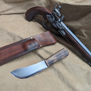 May include: A replica flintlock pistol with a dark wood grip and metal components, placed with a leather sheath and a knife with a wooden handle and curved blade. The items are displayed on a tan fabric background.