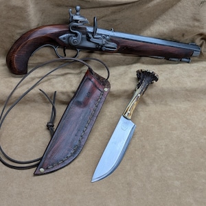 May include: A flintlock pistol with a dark wood stock and metal barrel, accompanied by a fixed-blade knife featuring an antler handle and a dark brown leather sheath with a long leather cord for carrying or attachment.