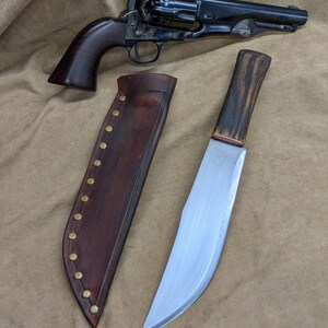May include: A black revolver with a wooden grip and a large, curved blade knife with a wooden handle and a leather sheath. The sheath is brown with brass rivets. The items are displayed on a beige surface.