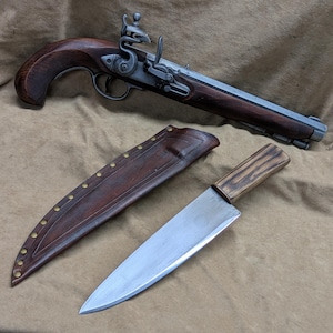 May include: A silver and brown flintlock pistol with a wooden handle and a leather sheath for a knife. The knife has a wooden handle and a silver blade.