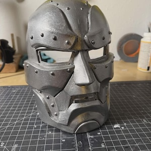 May include: A metallic silver full-face mask featuring a rigid, angular design with prominent brow ridges, a central nose bridge, and a segmented jawline. The surface is adorned with small, raised circular rivets along the edges and seams.