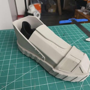 Puede incluir: Una carcasa de zapato rígida y estructurada de color gris claro con un diseño geométrico segmentado y una suela gruesa con relieve. El exterior presenta un acabado mate liso con líneas de panel visibles y un forro interior de tela negra.