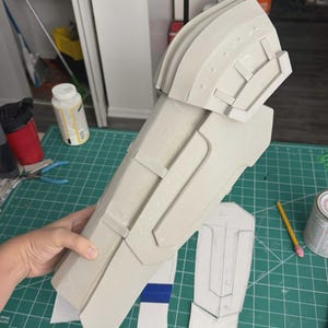 May include: A light gray foam armor gauntlet featuring a layered, segmented design with a rounded shoulder cap and raised rectangular panels. The piece has a smooth, matte finish and is constructed from multiple joined foam sections.