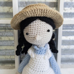 May include: A crocheted doll with black hair, wearing a white dress, a blue shirt, and a brown straw hat.