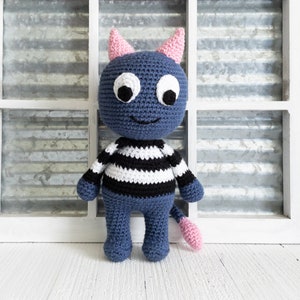 May include: A blue crocheted stuffed toy with pink ears and a tail. The toy has black and white striped clothing and large white eyes.