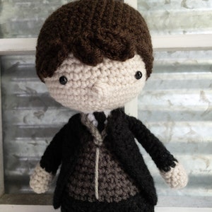 May include: A crocheted doll with brown hair, wearing a black suit with a brown vest. The doll has black eyes and a neutral expression.