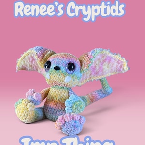 Puede incluir: Una criatura tejida a ganchillo con orejas grandes, una cola larga y dos colmillos blancos, con un patrón de hilo pastel multicolor. El texto Renee's Cryptids e Imp Thing aparece en azul claro con un borde blanco sobre un fondo rosa sólido.